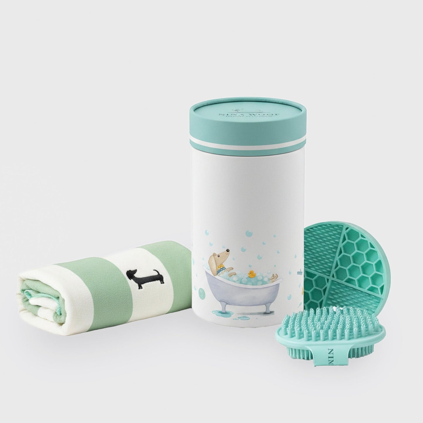 Vista aislada sobre fondo neutro del set de cuidado de baño para perros, mostrando la toalla enrollada, el envase cilíndrico decorado, la alfombrilla texturizada y el cepillo.
