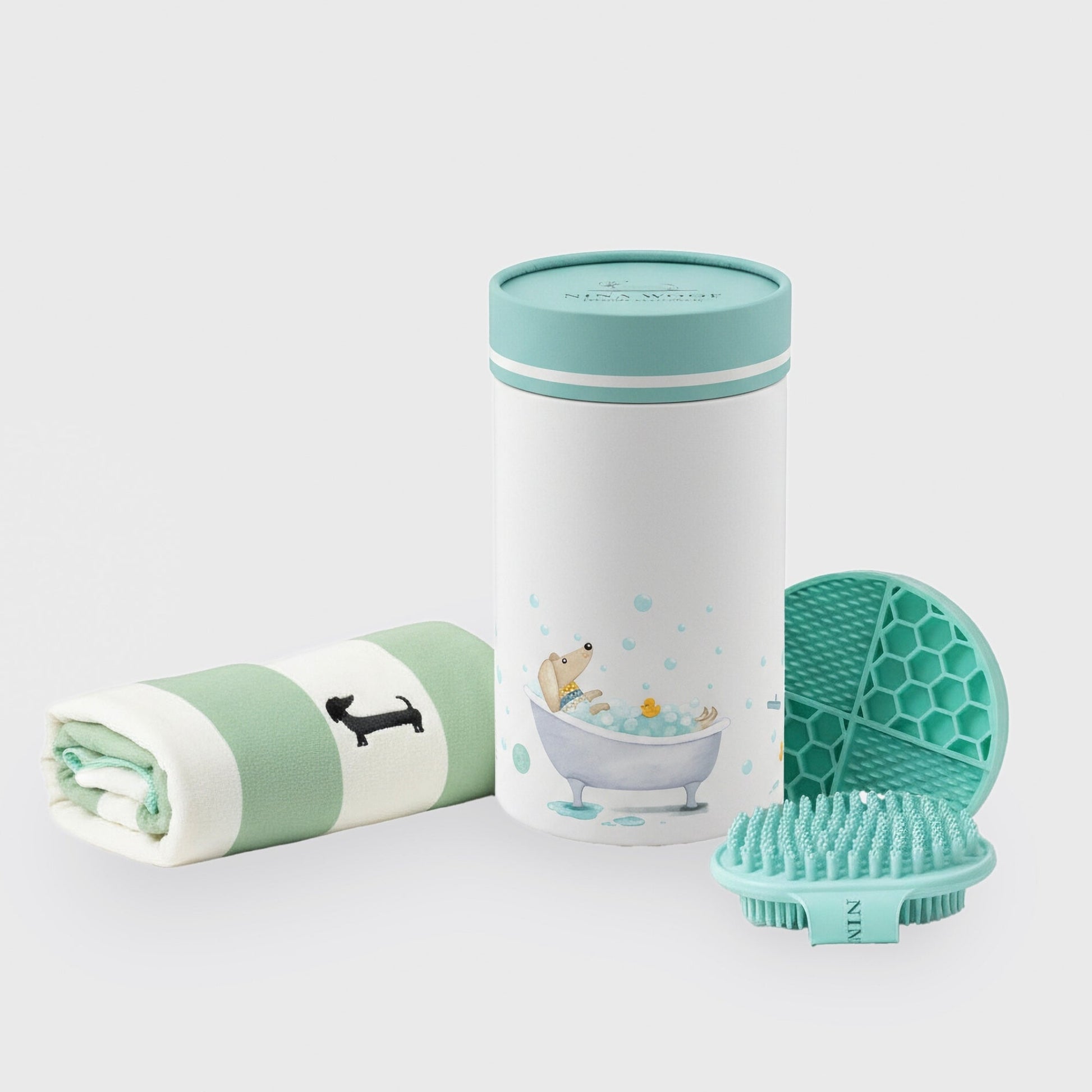 Vista aislada sobre fondo neutro del set de cuidado de baño para perros, mostrando la toalla enrollada, el envase cilíndrico decorado, la alfombrilla texturizada y el cepillo.